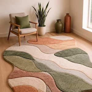 Puede incluir: Una alfombra de forma irregular con textura de mechones y bandas onduladas y orgánicas de colores verde salvia, naranja terracota, crema y beige. La alfombra tiene un pelo suave y una silueta fluida que no es rectangular.