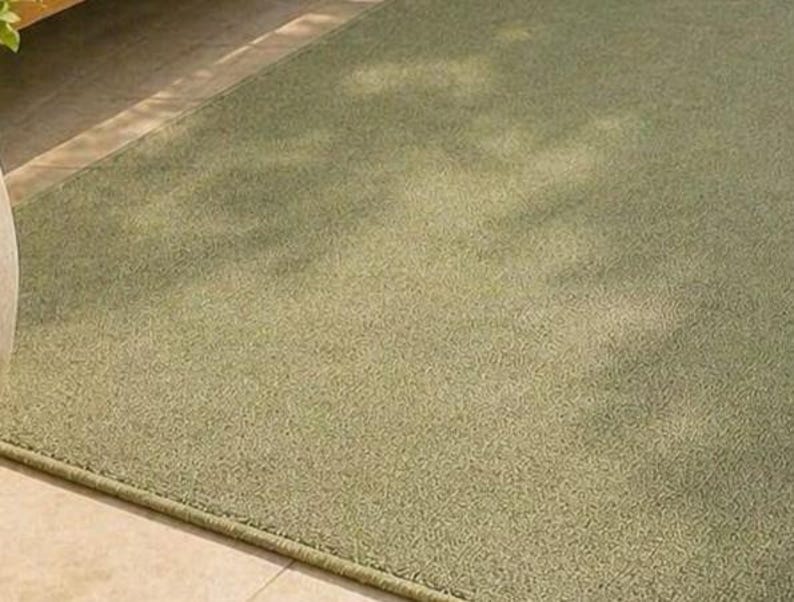 May include: A rectangular, sage green rug with a textured surface. The rug has a subtle, light-colored pattern and a darker green border. The rug is placed on a light-colored tile floor.