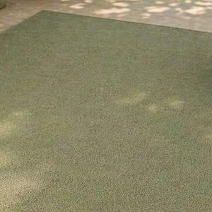 May include: A rectangular, olive green rug with a textured surface. The rug is placed on a light-colored concrete surface, with the legs of a wooden chair visible in the corners. The rug appears to be made of a durable material.