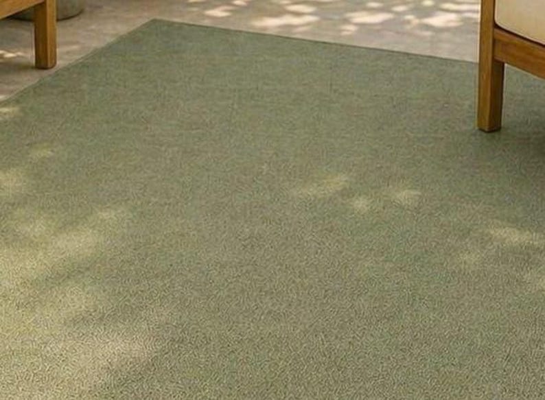 May include: A rectangular, olive green rug with a textured surface. The rug is placed on a light-colored concrete surface, with the legs of a wooden chair visible in the corners. The rug appears to be made of a durable material.