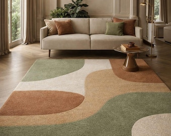 May include: A modern area rug with an abstract design in shades of green, brown, and cream. The rug is placed in a living room with a beige sofa, decorative pillows, and a glass-topped coffee table. The room has hardwood floors and a chandelier.