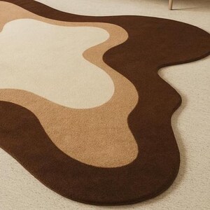 Puede incluir: Una alfombra moderna con un dise&ntilde;o abstracto. La alfombra presenta una forma fluida y org&aacute;nica con capas de color. Los colores incluyen tonos de marr&oacute;n, beige y crema, creando una est&eacute;tica c&aacute;lida. La alfombra est&aacute; hecha de un material suave y afelpado.