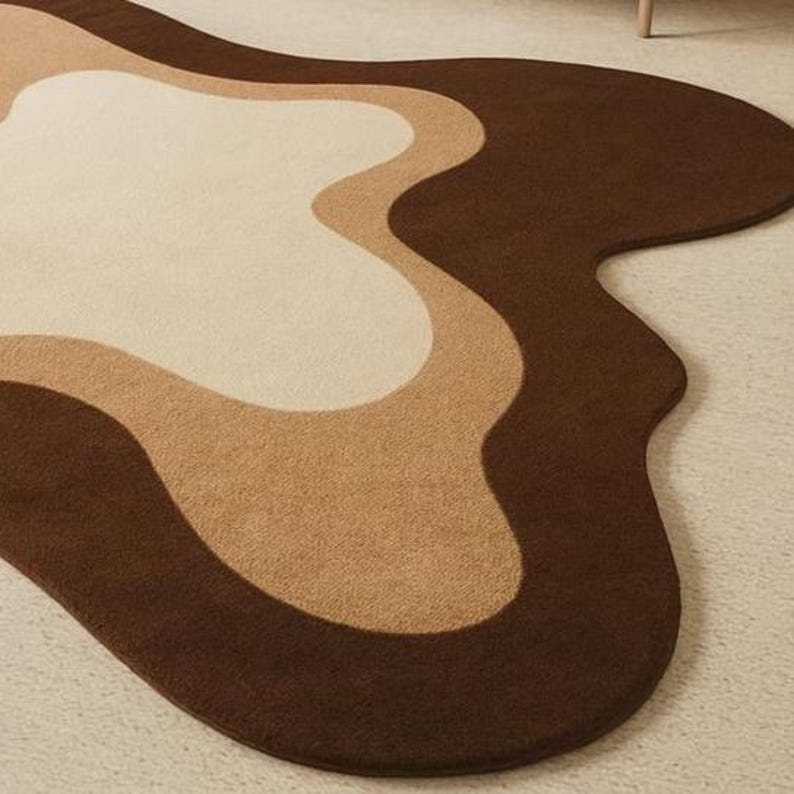 Puede incluir: Una alfombra moderna con un dise&ntilde;o abstracto. La alfombra presenta una forma fluida y org&aacute;nica con capas de color. Los colores incluyen tonos de marr&oacute;n, beige y crema, creando una est&eacute;tica c&aacute;lida. La alfombra est&aacute; hecha de un material suave y afelpado.