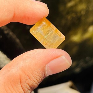 Puede incluir: Una gema rectangular de color amarillo dorado con inclusiones internas en forma de cabello. La gema se sostiene entre dos dedos, mostrando sus facetas pulidas y su calidad translúcida. El color de la piedra varía de amarillo claro a oscuro.