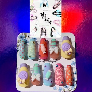 Peut inclure: Un ensemble de faux ongles avec divers motifs, dont des pois, des spirales, des bonbons et des nœuds. Les ongles sont dans des tons de bleu, orange, rose et jaune. Une carte avec le texte "THIS IS FOR" est en arrière-plan.