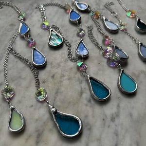 May include: A selection of handmade stained glass pendants in shades of blue, green, and teal. Each teardrop-shaped pendant is framed in silver and suspended on a silver chain. Iridescent beads add a touch of sparkle to the necklaces.