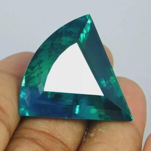 May include: A loose, faceted gemstone with a triangular shape and curved edges. The stone displays a vibrant teal and blue color gradient with high clarity and a polished surface that reflects light across its multiple geometric facets.