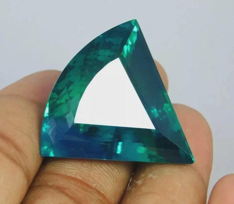 May include: A loose, faceted gemstone with a triangular shape and curved edges. The stone displays a vibrant teal and blue color gradient with high clarity and a polished surface that reflects light across its multiple geometric facets.