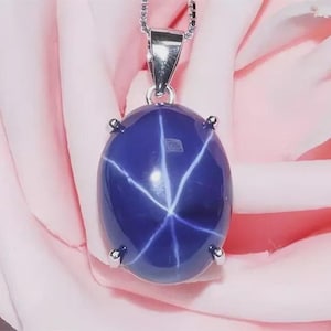 以下が含まれることがあります： 星のような模様が特徴的な、大きな楕円形の濃い青色の宝石が付いたペンダントネックレス。 宝石は銀色の金属製のセッティングにセットされ、繊細なチェーンから吊り下げられています。 背景はソフトピンクです。