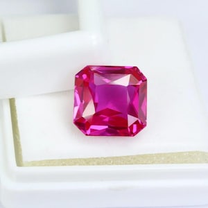 May include: A vibrant, rectangular-cut, hot pink gemstone. The gem is set against a white background, showcasing its facets and internal reflections. The stone is a deep, saturated pink, with a polished, reflective surface.