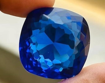 May include: A large, cushion-cut gemstone in a deep, vibrant blue hue. The gem is held between two fingers, showcasing its intricate internal reflections and brilliant facets. The overall shape is a rounded square, with a polished surface.