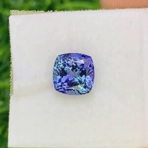 Pu&ograve; includere: Una gemma tagliata quadrata, multifattoriale, con una miscela di tonalit&agrave; blu e viola. La gemma &egrave; posta su uno sfondo bianco, evidenziando il suo colore e la sua chiarezza. Le sfaccettature della pietra riflettono la luce.
