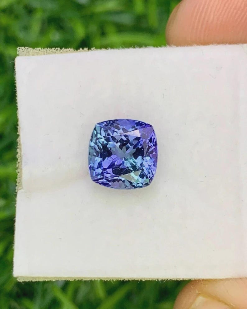 Pu&ograve; includere: Una gemma tagliata quadrata, multifattoriale, con una miscela di tonalit&agrave; blu e viola. La gemma &egrave; posta su uno sfondo bianco, evidenziando il suo colore e la sua chiarezza. Le sfaccettature della pietra riflettono la luce.