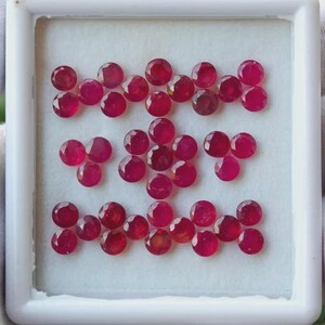 May include: A selection of round, faceted ruby gemstones in a white tray. The rubies are a deep red colour and arranged in a grid pattern. The gemstones are likely for jewellery making or collecting.