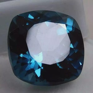 May include: A cushion-cut gemstone with a deep teal color and a bright, white center. The facets of the gem reflect light, creating a sparkling effect. The gem is set against a neutral background.