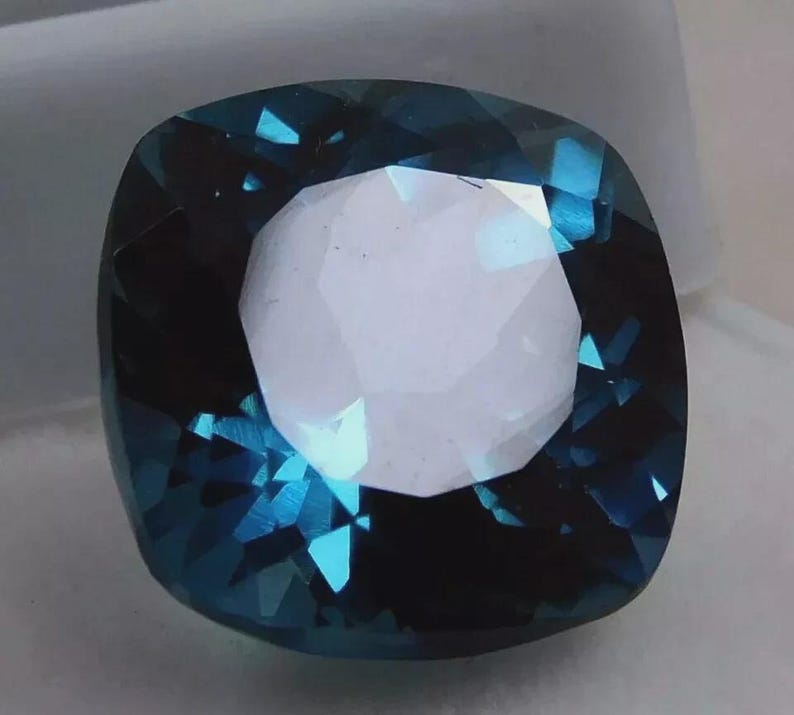 May include: A cushion-cut gemstone with a deep teal color and a bright, white center. The facets of the gem reflect light, creating a sparkling effect. The gem is set against a neutral background.
