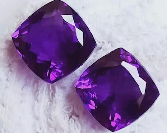 May include: Two cushion-cut amethyst gemstones. The gems are a deep purple colour with facets that reflect light. The stones are set against a white, textured background.