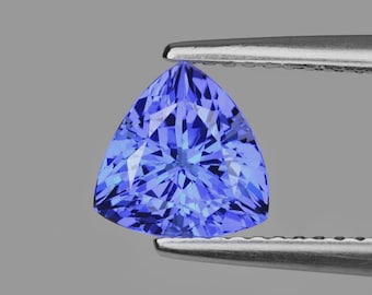 May include: A triangular, blue gemstone with multiple facets, held by silver tweezers. The gem's cut creates a brilliant display of light. The background is a plain, neutral grey.