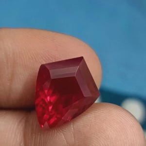 Op de afbeelding: Een gefacetteerde, dieprode edelsteen met een unieke geometrische vorm. De edelsteen heeft meerdere facetten die licht reflecteren, waardoor een briljante glinstering ontstaat. De steen wordt tegen een vage blauwe achtergrond gehouden.