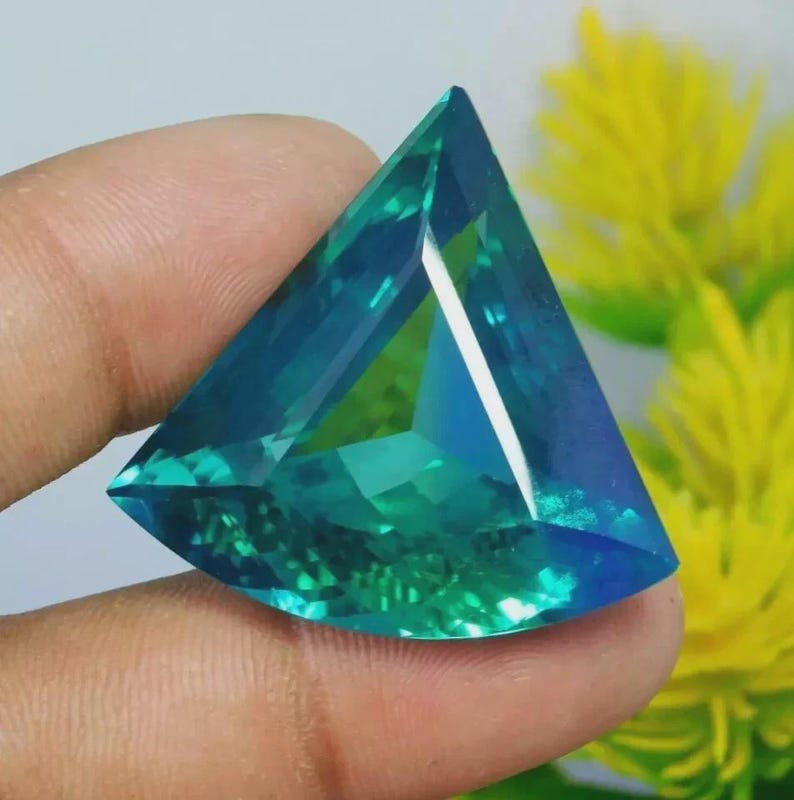 May include: A triangular faceted gemstone displaying a vibrant gradient of deep blue and bright green hues. The stone features sharp, clean edges and a polished surface that reflects light, creating a clear, transparent appearance with internal depth.