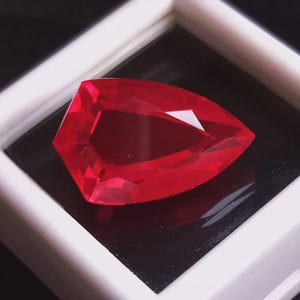 May include: A faceted, vibrant red gemstone with a unique, angular cut. The gem is displayed in a white-framed box against a black background, highlighting its brilliance and color. The shape is a modified shield or kite.