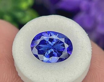May include: A deep blue, oval-cut gemstone with multiple facets, set on a white circular base. The background is blurred, with purple and green hues. The gemstone's facets reflect light, creating a sparkling effect. Likely a Tanzanite gemstone.