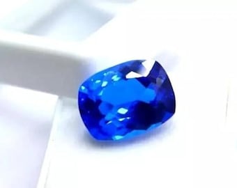 May include: A brilliant blue, cushion-cut gemstone. The facets of the gem catch the light, creating a sparkling effect. The gem is displayed against a white backdrop, accentuating its colour and clarity. The gem is likely a sapphire.