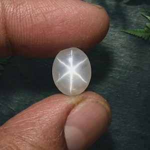 May include: A translucent, oval-shaped cabochon gemstone featuring a distinct six-rayed star effect known as asterism. The stone displays a soft, milky white color with a centered, bright white star pattern that reflects light across its smooth surface.