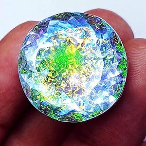 May include: A large, round gemstone with a faceted surface. The gem features a vibrant display of colors, including blues, greens, and yellows, creating a shimmering effect. The gemstone is held in a hand, showcasing its size and brilliance.