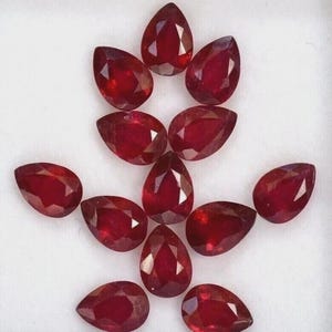 May include: A collection of eleven pear-shaped, faceted rubies. The gemstones are a deep, rich red color and arranged in a circular pattern against a white background. Each stone has multiple facets that catch the light.