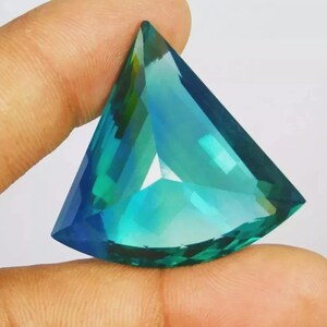 May include: A triangular-cut gemstone featuring a vibrant gradient of teal, blue, and green hues. The stone has a faceted surface that reflects light, creating a translucent appearance with sharp edges and a pointed top.
