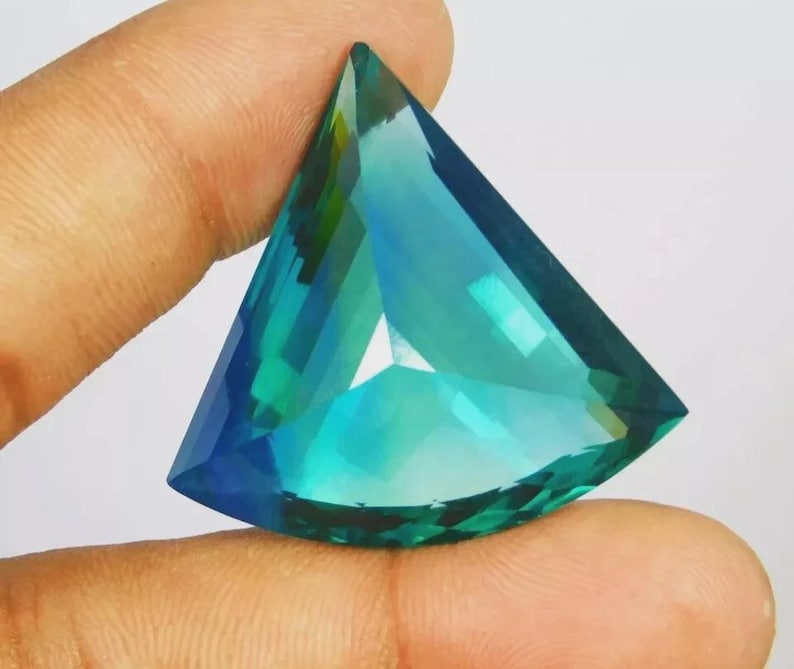 May include: A triangular-cut gemstone featuring a vibrant gradient of teal, blue, and green hues. The stone has a faceted surface that reflects light, creating a translucent appearance with sharp edges and a pointed top.