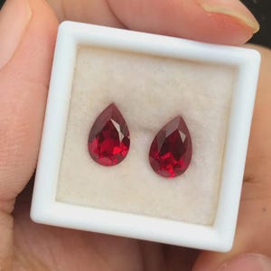 Könnte beinhalten: Zwei facettierte, birnenförmige, tiefrote Edelsteine werden in einer weißen Box präsentiert. Die Edelsteine haben einen Brillantschliff, der das Licht reflektiert. Die Box wird von einer Hand gehalten, die die Farbe und Klarheit der Edelsteine zeigt. Diese Edelsteine eignen sich für die Schmuckherstellung.