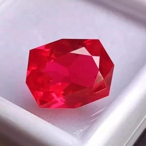 May include: A faceted, vibrant red gemstone with a complex, geometric cut. The gem is set against a white background, highlighting its brilliant, reflective surfaces. The gemstone's color is a deep, saturated red.