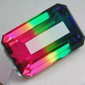 May include: A rectangular gemstone with a vibrant array of colors, including pink, green, yellow, and blue. The gem has a large, white, rectangular facet in the center. The gemstone is cut with multiple facets, creating a sparkling effect.