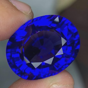 May include: A faceted, oval-shaped, deep blue gemstone. The gem is held between fingers, showcasing its brilliant cut and internal facets. The stone's color ranges from a vibrant, saturated blue to darker shades within.