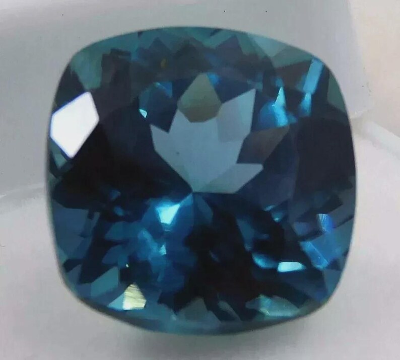 May include: A faceted, cushion-cut gemstone with a deep teal color. The gem's surface reflects light, creating a sparkling effect. The stone's shape is square with rounded corners, and it appears to be set against a white background.