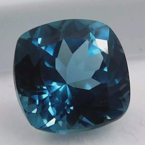 May include: A faceted, cushion-cut gemstone with a deep teal color. The gem's surface reflects light, showcasing its intricate internal structure and facets. The overall shape is square with rounded corners, typical of a cushion cut.