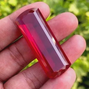 May include: A faceted, rectangular gemstone with a deep red hue. The gem is held in a hand, showcasing its polished surfaces and internal clarity. The cut of the stone creates multiple reflective facets.