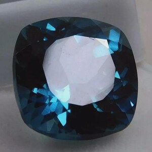 May include: A cushion-cut gemstone with a deep teal color and a bright, white center. The facets of the gem reflect light, creating a sparkling effect. The gem is set against a neutral background.