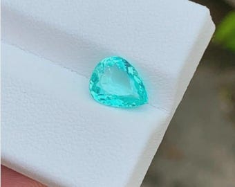 May include: A pear-shaped, light teal gemstone. The gem is set against a white background. The gemstone has a transparent appearance and a polished surface, reflecting light. The overall shape is a teardrop, with a rounded top and a pointed bottom.