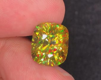 May include: A cushion-cut gemstone with a vibrant yellow hue, displaying internal reflections of green and red. The gemstone is held between fingers against a dark background, showcasing its facets and brilliance.