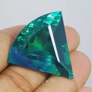 May include: A faceted gemstone with a fan-like shape, displaying a gradient of teal and blue hues. The gem's surface reflects light, showcasing its cut and clarity. The gemstone is held in a hand.