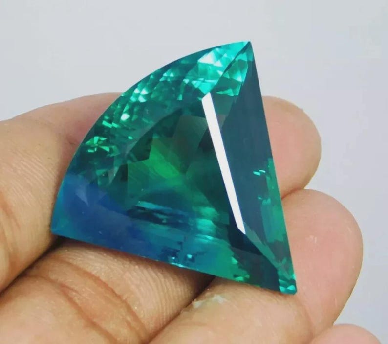 May include: A faceted gemstone with a fan-like shape, displaying a gradient of teal and blue hues. The gem's surface reflects light, showcasing its cut and clarity. The gemstone is held in a hand.