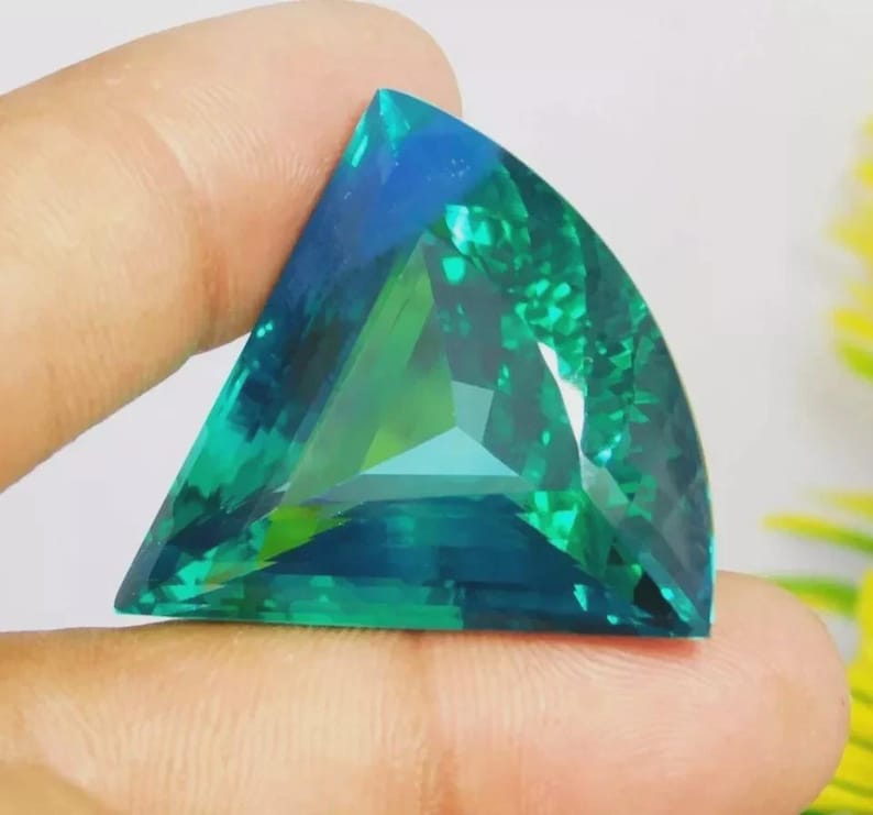 May include: A triangular-cut gemstone featuring a vibrant gradient of deep blue and emerald green hues. The faceted surface displays a complex internal pattern with light reflecting across the polished edges of the stone.