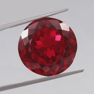 May include: A round, faceted, deep red gemstone held by silver tweezers. The gem's multiple surfaces reflect light, creating a brilliant display. The gemstone is a vibrant, rich red colour.