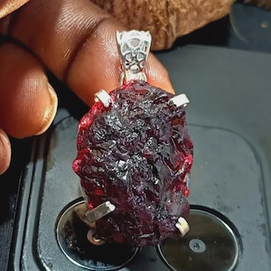 Puede incluir: Un colgante de granate en bruto, de color rojo oscuro, con una fianza y puntas de color plateado. La piedra preciosa tiene una superficie rugosa y texturizada y está suspendida de una fianza decorativa. El colgante es una piedra preciosa natural sin pulir.