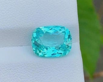 May include: A vibrant, cushion-cut gemstone in a bright turquoise hue. The gem is set against a white background, with a glimpse of green foliage on the right. The facets of the stone catch the light, creating a sparkling effect.