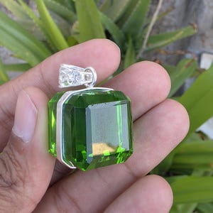 May include: A faceted, emerald-green gemstone pendant with a silver-colored bail. The rectangular-cut stone is set in a silver frame. The pendant is held up against a background of green foliage.