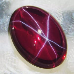 May include: Oval-shaped, deep red gemstone with a six-rayed star pattern. The polished surface reflects light, creating a vibrant, eye-catching display. The gemstone's color is a rich, saturated red, with a smooth, glossy finish.